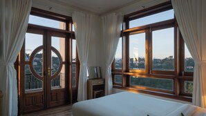Habitació d'hotel amb un llit gran, terra de fusta i vista a la ciutat a través de grans finestres amb cortines.