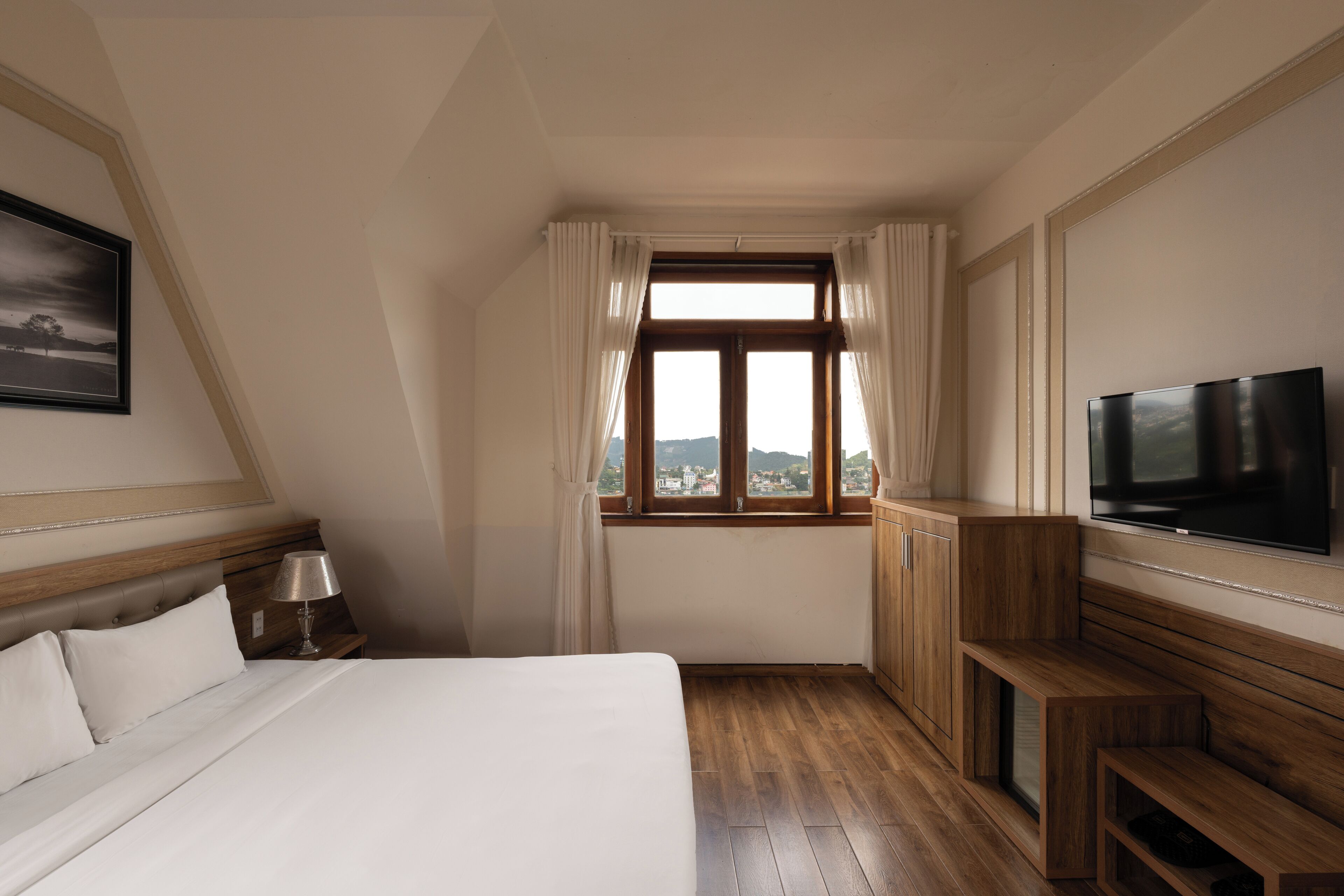 A hotel room with a bed, a TV, a wooden bench, a nightstand, and a window with curtains.