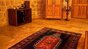 A stone-walled room with a flat-screen TV mounted on the wall, a wooden cabinet, a decorative floor rug, and a wooden door. - Songül Hanım Konağı (Ürgüp)