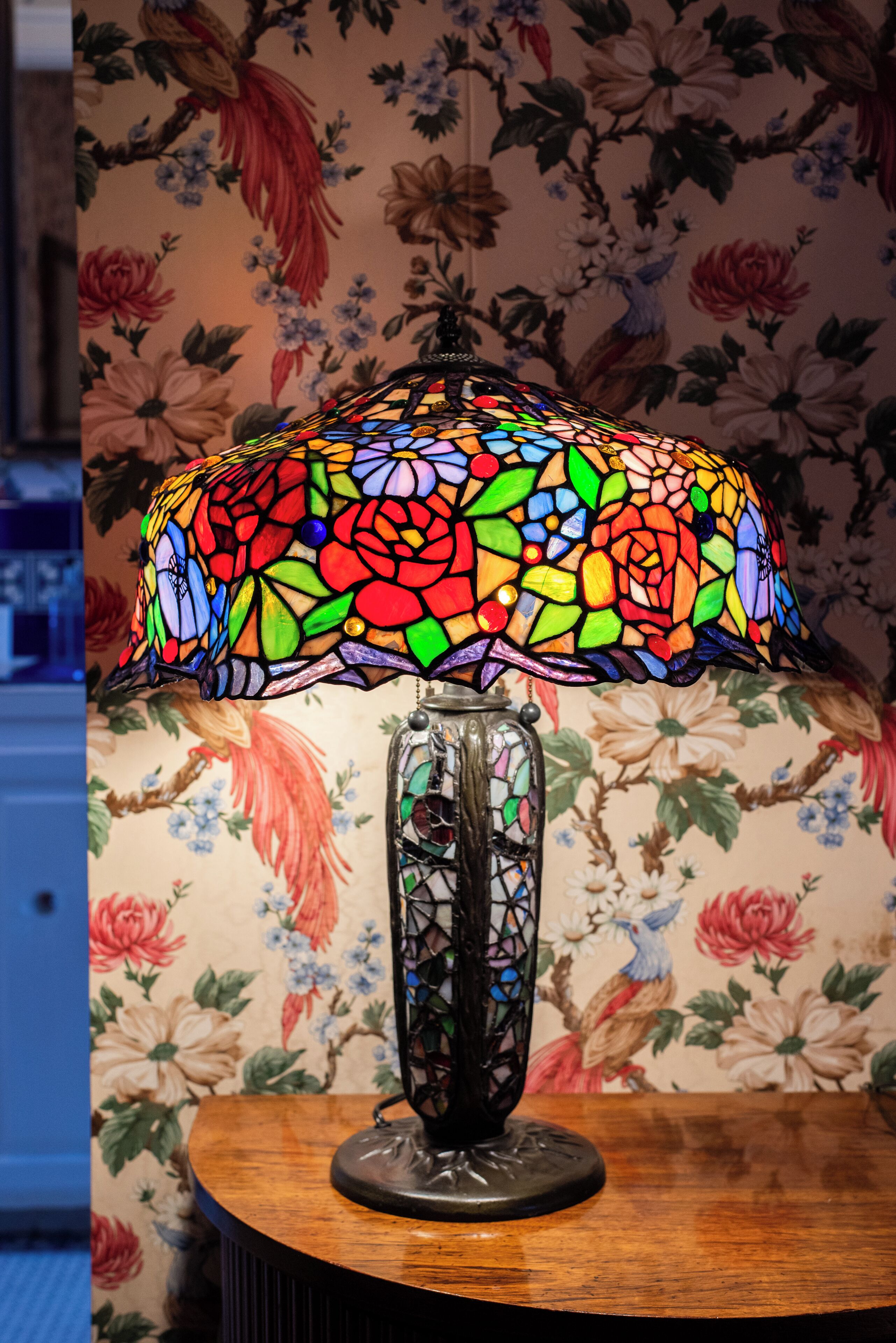 En bordlampe med skærm i farvet glas og et dekorativt stel, stående på et træbord foran blomstret tapet.