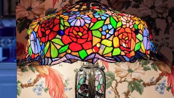En bordlampe med skærm i farvet glas og et dekorativt stel, stående på et træbord foran blomstret tapet.