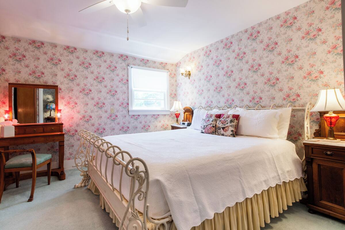 A bedroom with floral wallpaper, a white bed, a wooden desk, and a nightstand with a lamp.