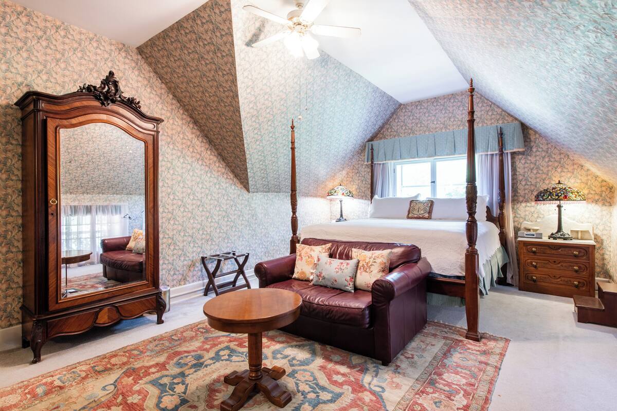 A bedroom with a four-poster bed, a large mirror, a leather sofa, and a small round table.