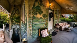A covered porch with a mural, outdoor seating, and a view of a garden.