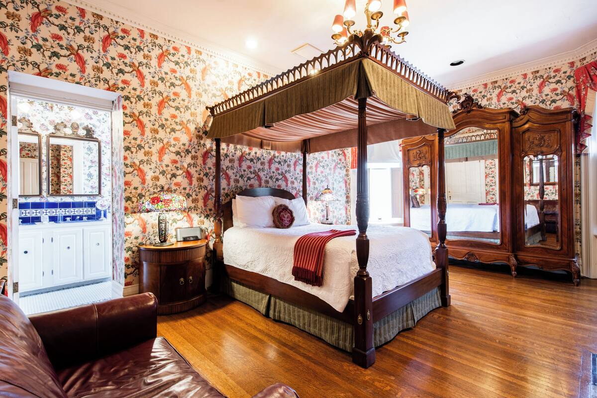 A four-poster canopy bed with a wooden frame and a red bedspread, a white wardrobe with a mirror, and floral wallpaper.