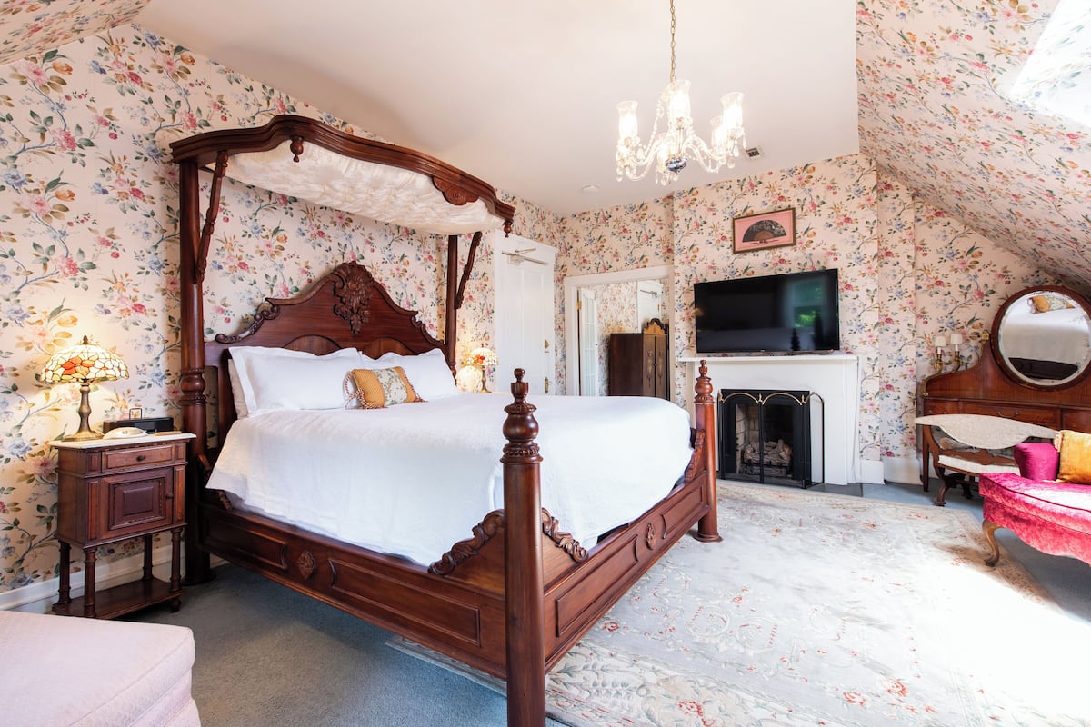 A bedroom with a four-poster bed, a chandelier, a television, and a fireplace.