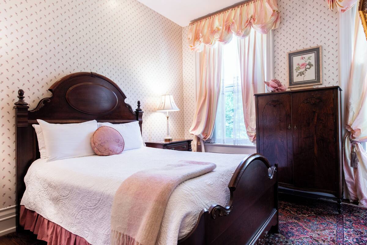 A neatly made bed with a headboard, a bedside table with a lamp, a wooden wardrobe, and a window with curtains.