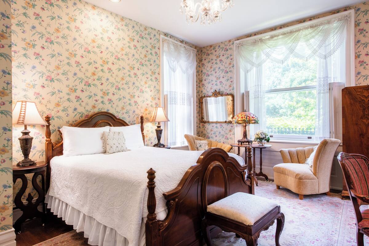 A bedroom with floral wallpaper, a wooden bed, two bedside tables with lamps, a chair, and a mirror.