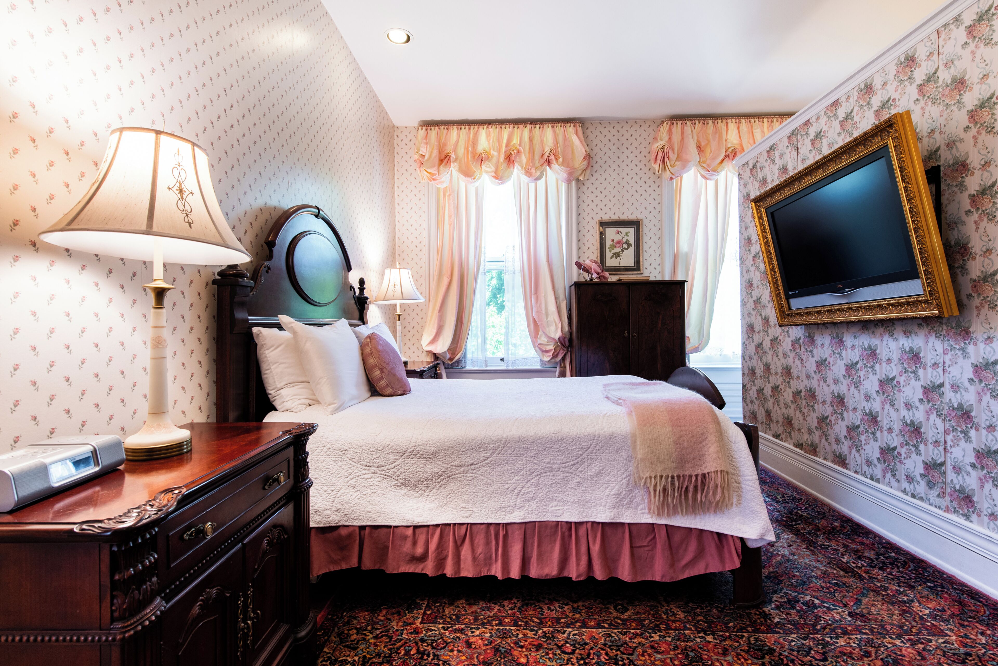 A bedroom with a bed, two bedside lamps, a dresser, a television mounted on the wall, and a window with curtains.