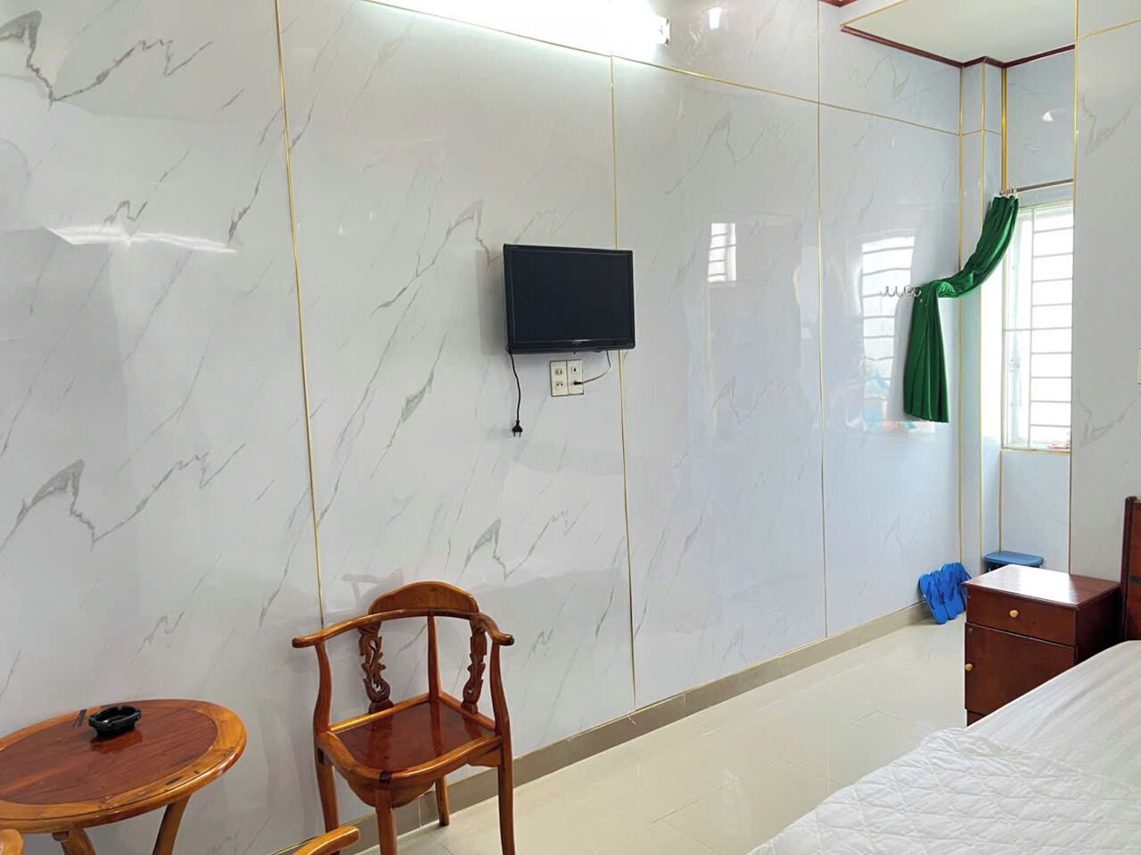 Een hotelkamer met een bed, een houten stoel, een klein rond tafeltje, een aan de muur bevestigde televisie en een raam met een groene gordijn.