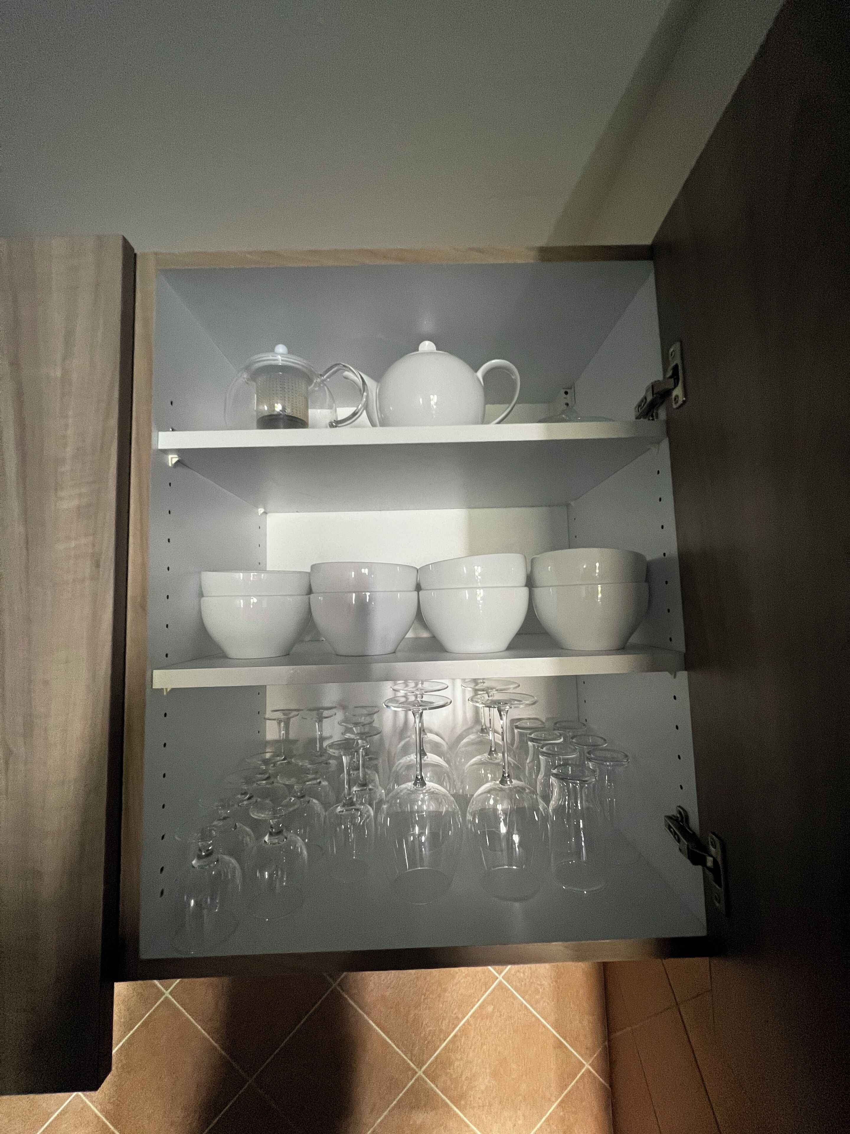 A kitchen cabinet with shelves holding white bowls, a teapot, and wine glasses.