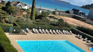 Une piscine rectangulaire à l’eau d’un bleu limpide, entourée d’une plage de sable. Des chaises longues sont alignées le long du bord de la piscine, et on aperçoit un petit plongeoir. La piscine est située devant un paysage de plage et d’océan, avec des bâtiments et de la végétation en arrière-plan.