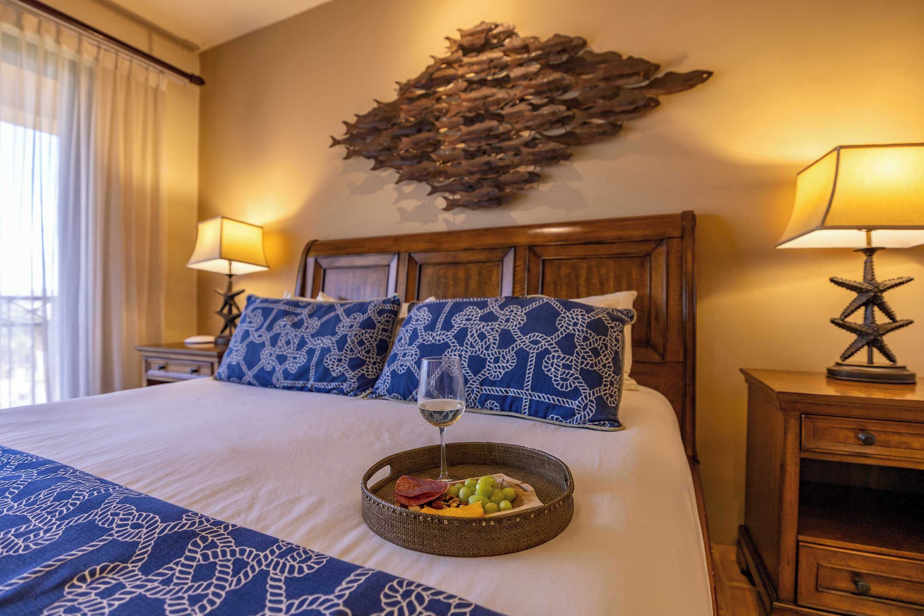 A hotel room with a bed, two bedside lamps, a decorative wall piece, and a tray with fruits and a glass of wine.