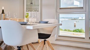 A dining area with a table set for two, a wine bottle, and glasses. A window offers a view of a body of water and greenery.