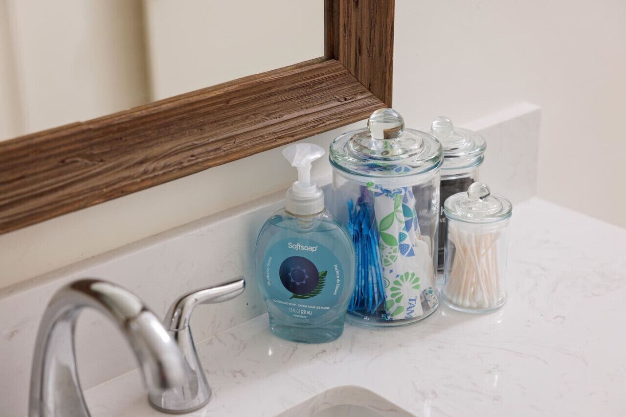 A bathroom sink with a mirror, a bottle of soft soap, and two glass jars with cotton swabs.