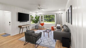 A modern living room with a flat-screen TV, grey sofas, a coffee table, and a large window offering a view of the outdoors. - Renovated Capital Heights Home (Baton Rouge)