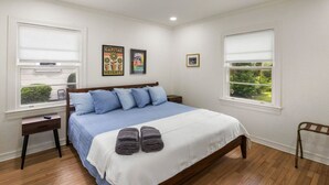 A bedroom with a large bed, two bedside tables, a bench, and two windows with blinds. - Renovated Capital Heights Home (Baton Rouge)