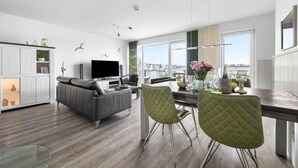 Una sala de estar moderna con un sofá gris, una mesa de comedor con sillas, un televisor y vistas de la ciudad a través de ventanas grandes.