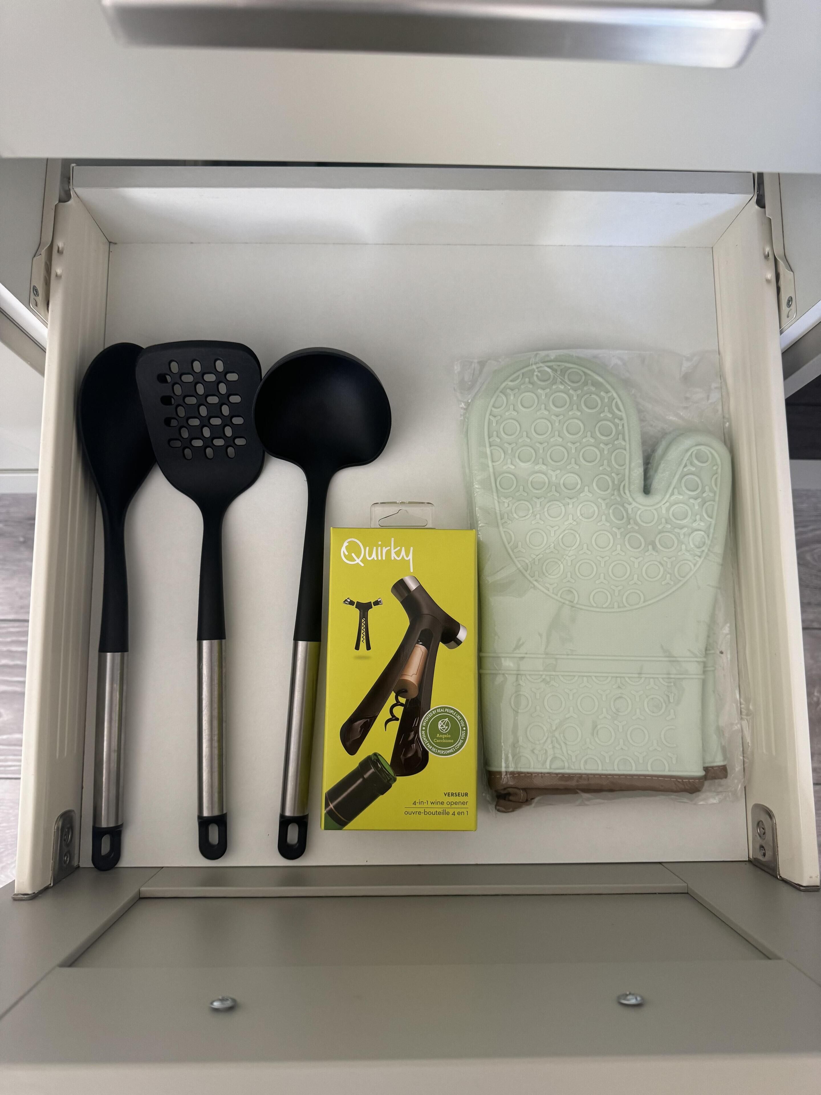Un cajón de cocina con una espátula negra, una cuchara de servir negra, una caja amarilla con la etiqueta 'Quirley' y un sacacorchos, además de un guante de horno de silicona verde.