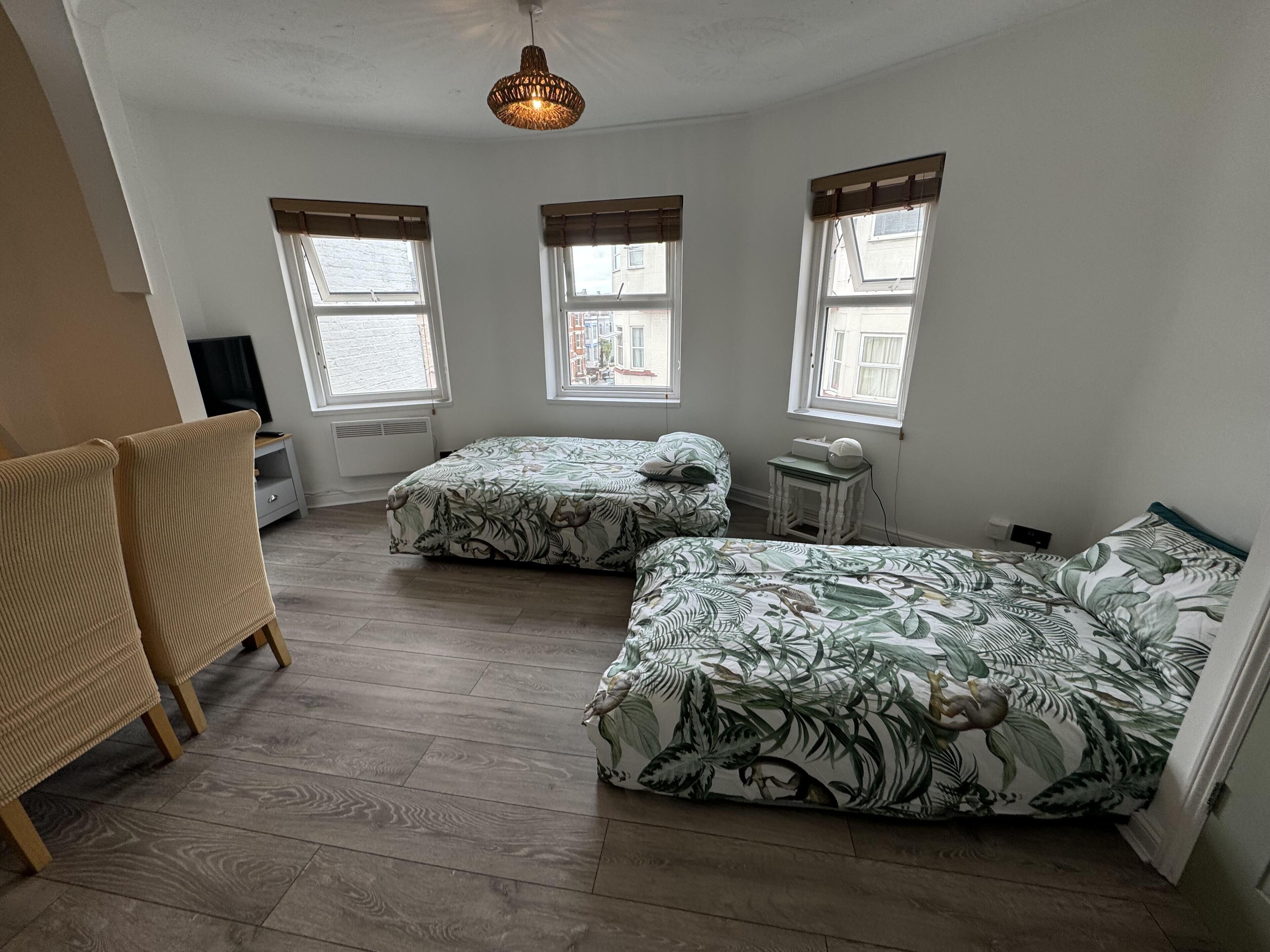 A room with two beds, a desk, and a chair. There are three windows with curtains.