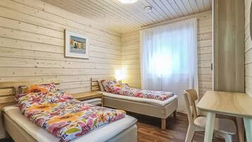 A room with two beds, a chair, a table, a picture on the wall, and a window with curtains.