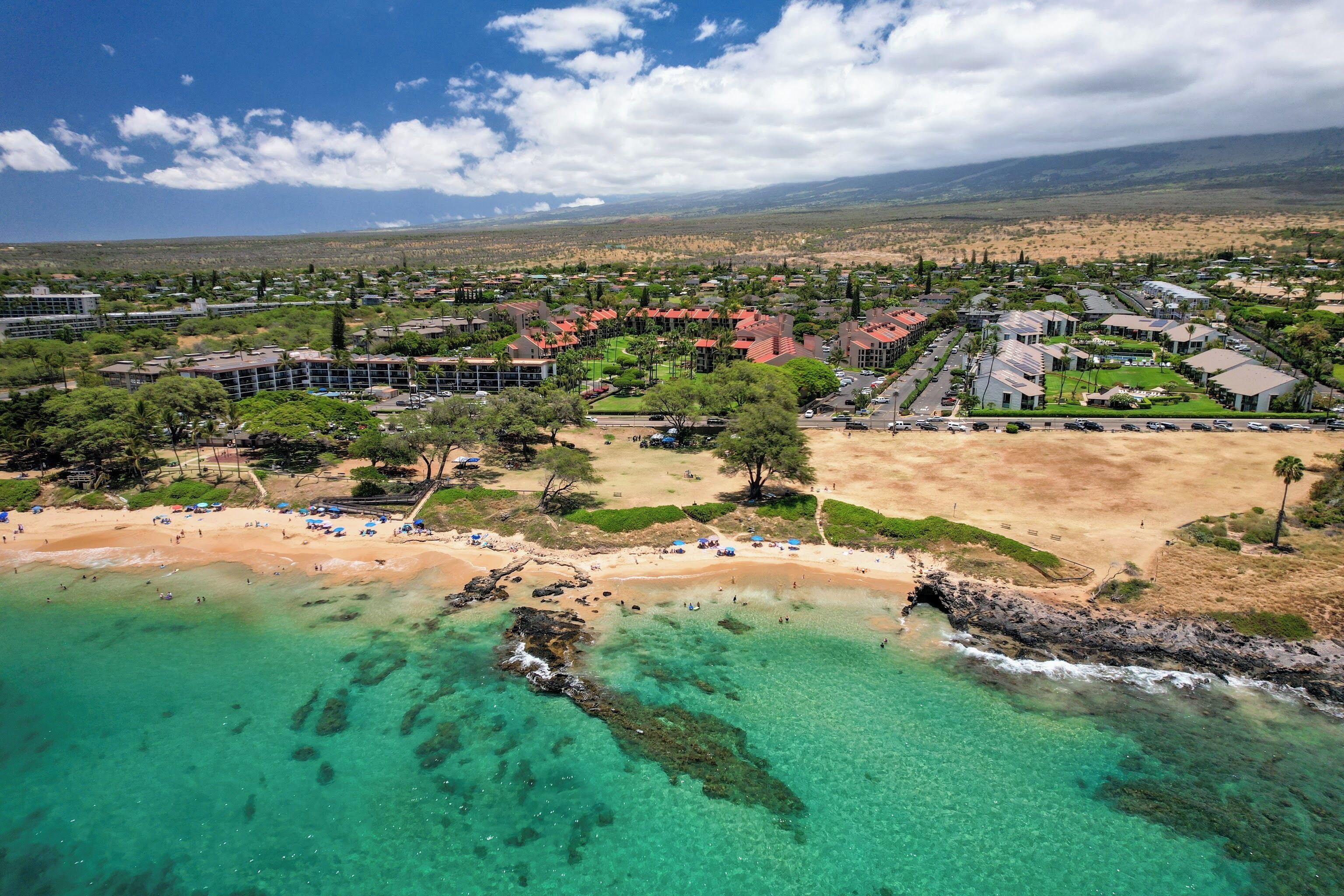 An aerial view of a coastal resort with a beach, clear turquoise waters, and a mountainous backdrop.