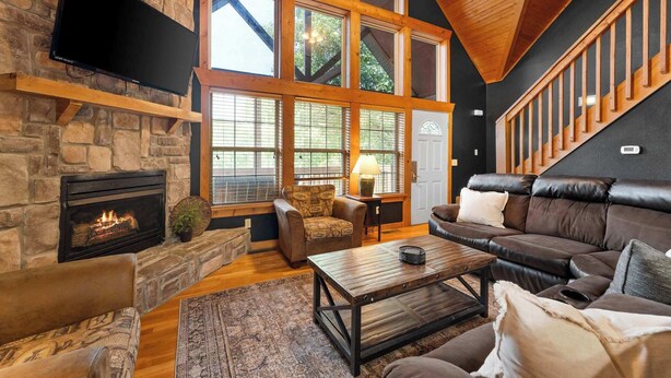 A cozy living room with a stone fireplace, leather sofa, and a large window with a view of trees. - Rustic Cabin with 4 Master Suites by SDC! (Branson West)