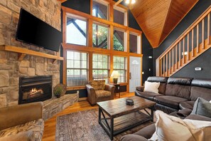 A cozy living room with a stone fireplace, leather sofa, and a large window with a view of trees. - Rustic Cabin with 4 Master Suites by SDC! (Branson West)