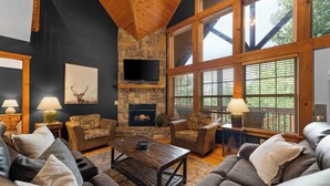 A cozy living room with a stone fireplace, leather sofas, and large windows. - Rustic Cabin with 4 Master Suites by SDC! (Branson West)