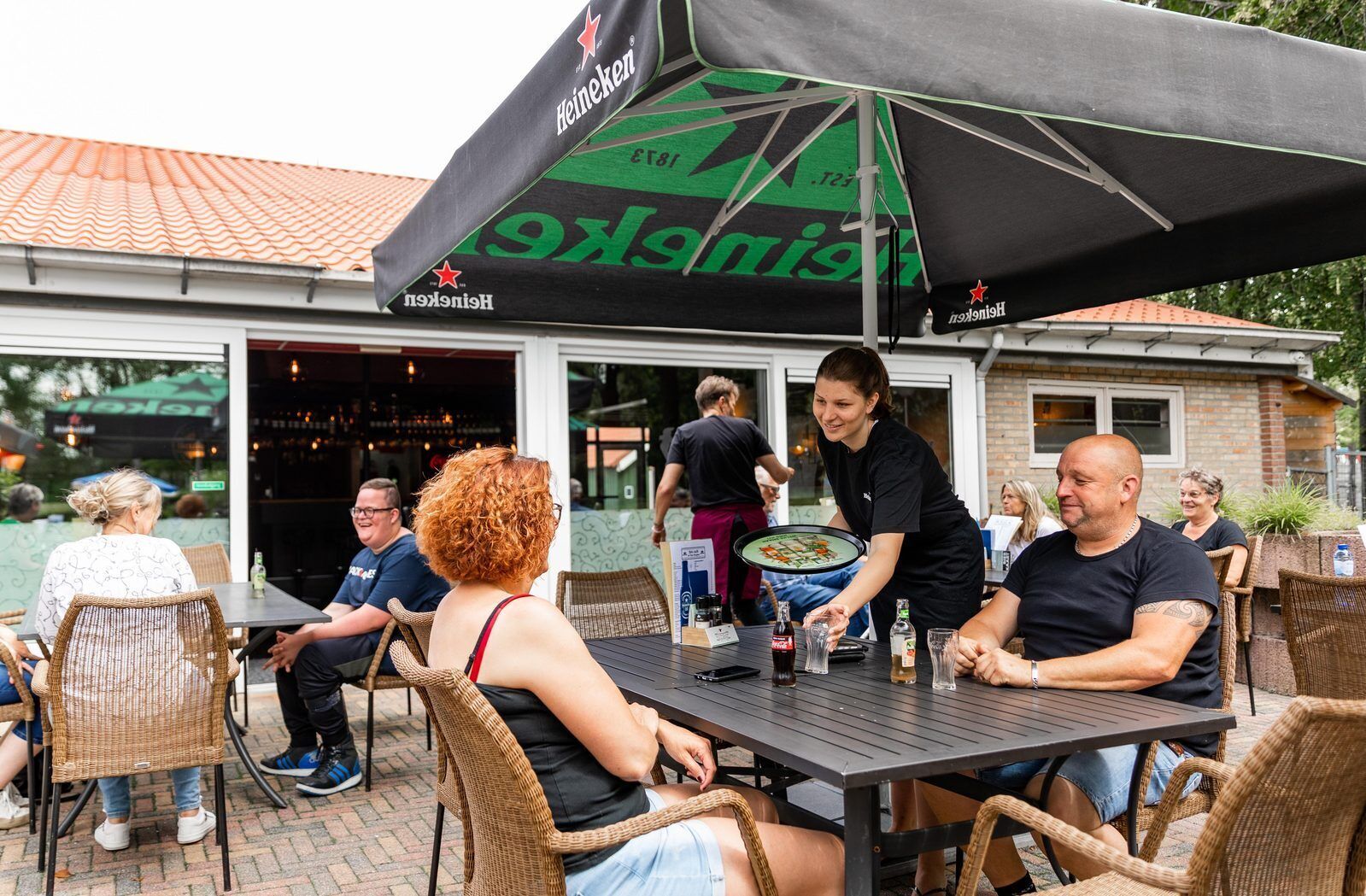 Ein Sitzbereich im Freien mit Korbstühlen und Tischen, einem mit dem Heineken-Logo versehenen Sonnenschirm und einem Backsteinhaus im Hintergrund.