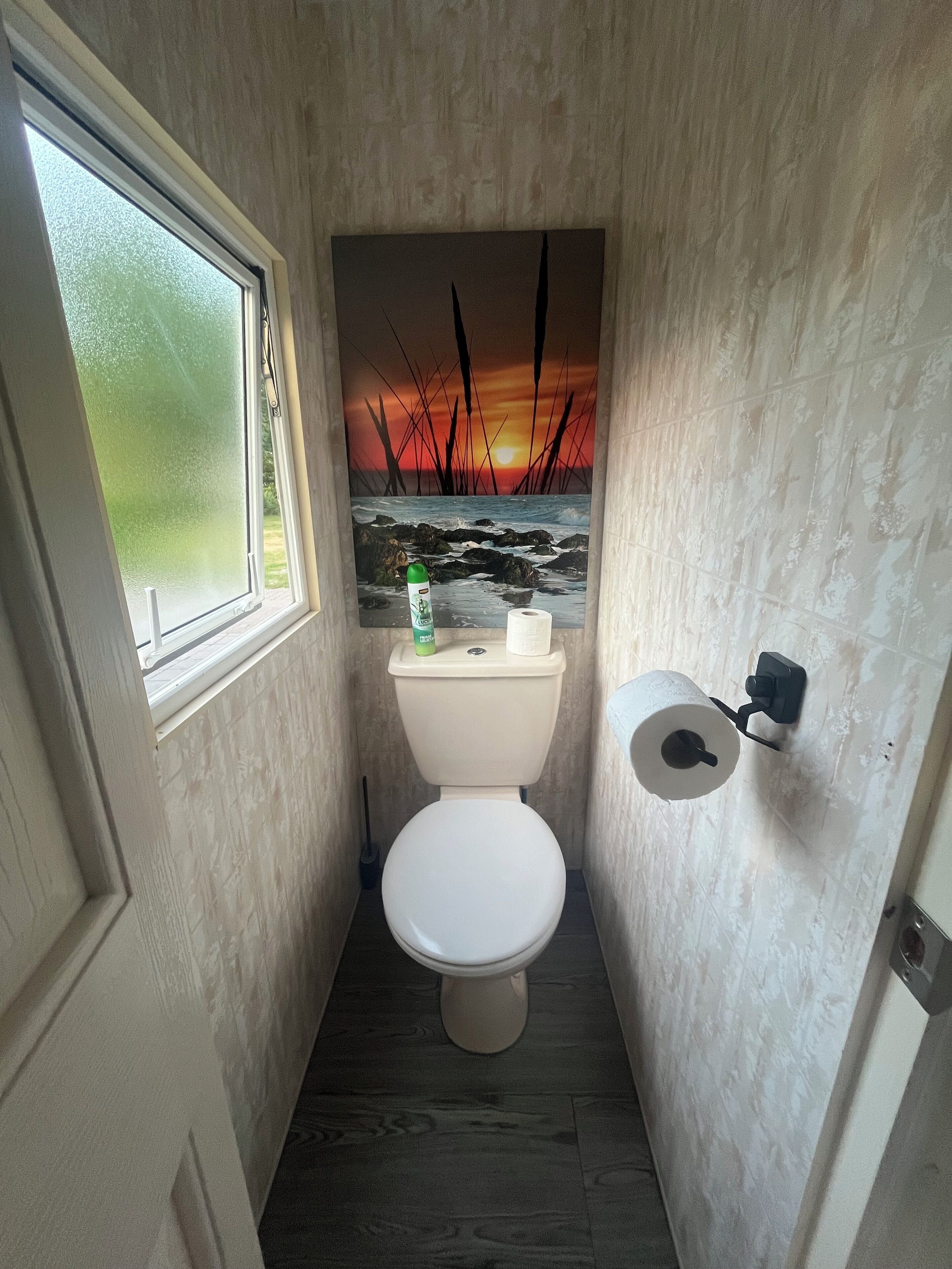 Et lille badeværelse med toilet, et vindue, et maleri af en solnedgang og en toiletpapirholder.