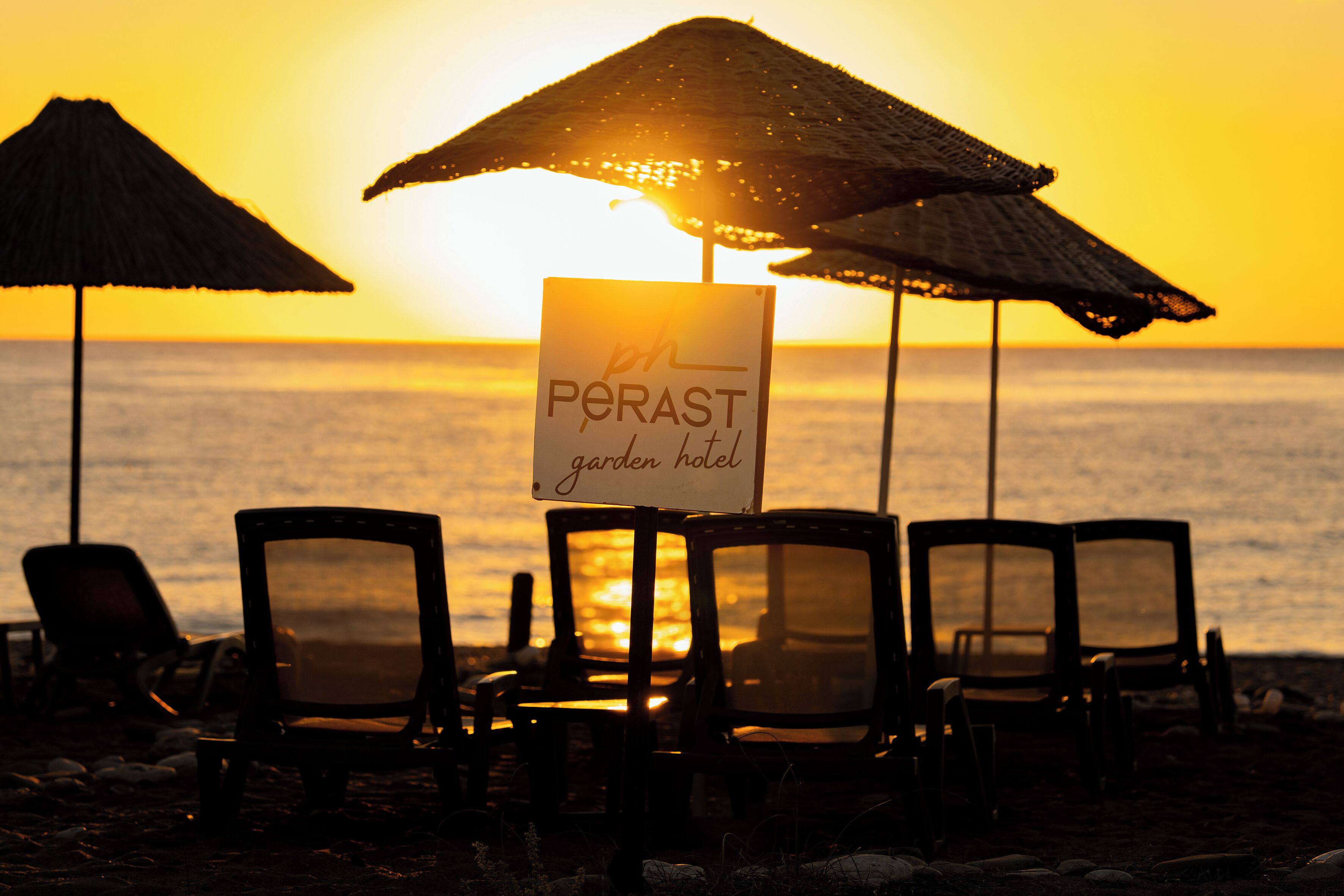 A beach scene with a sign for 'Perast Garden Hotel' and several beach chairs under umbrellas.