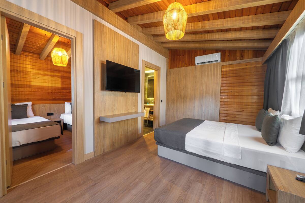 Una habitación de hotel moderna con paredes de madera, un televisor de pantalla plana, una cama con cubrecama gris y blanco, y un techo de madera con vigas a la vista.