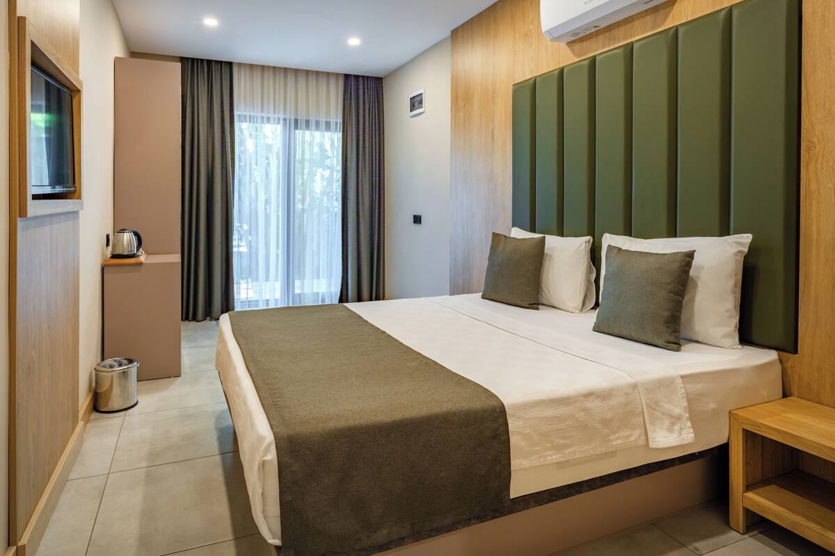 Una habitación de hotel moderna con una cama grande, un cabecero de madera, una mesita de noche pequeña y vistas al exterior a través de la ventana.