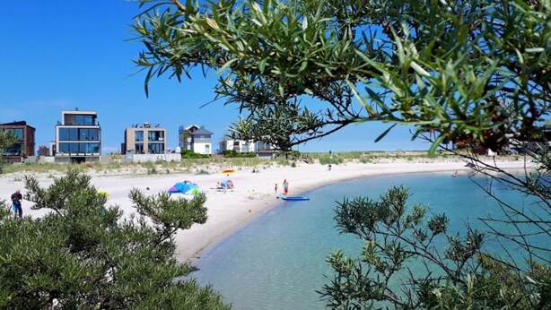 Ein Strand mit modernen Gebäuden im Hintergrund, klarem Himmel und ruhiger See.