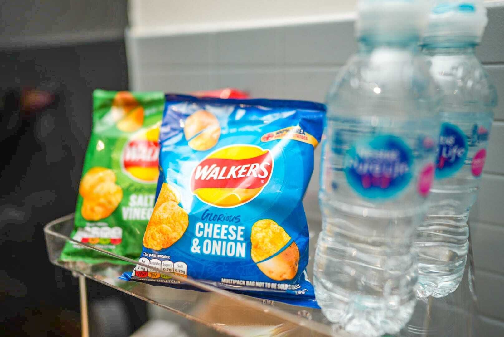 Un petit paquet de chips saveur fromage et oignon de la marque Walkers et deux bouteilles d’eau, posés sur une étagère.