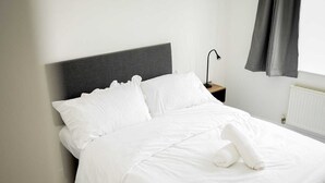 A neatly made bed with white linens and pillows, a dark headboard, a bedside table with a lamp, and a window with curtains. - One Bedroom Flat in Gravesend Kent (Gravesend)