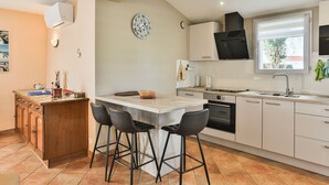 A modern kitchen with a central island, bar stools, a built-in oven, and a window with a view. - Côte Vendéenne Résidence le pin Parasol (Longeville-sur-Mer)