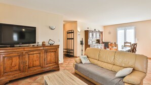 A living room with a TV, wooden cabinet, leather sofa, and a dining area with chairs and a table. - Côte Vendéenne Résidence le pin Parasol (Longeville-sur-Mer)