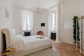 A bedroom with a large bed, a bedside table, a chair, a radiator, a window with curtains, a hanging light fixture, and a plant.