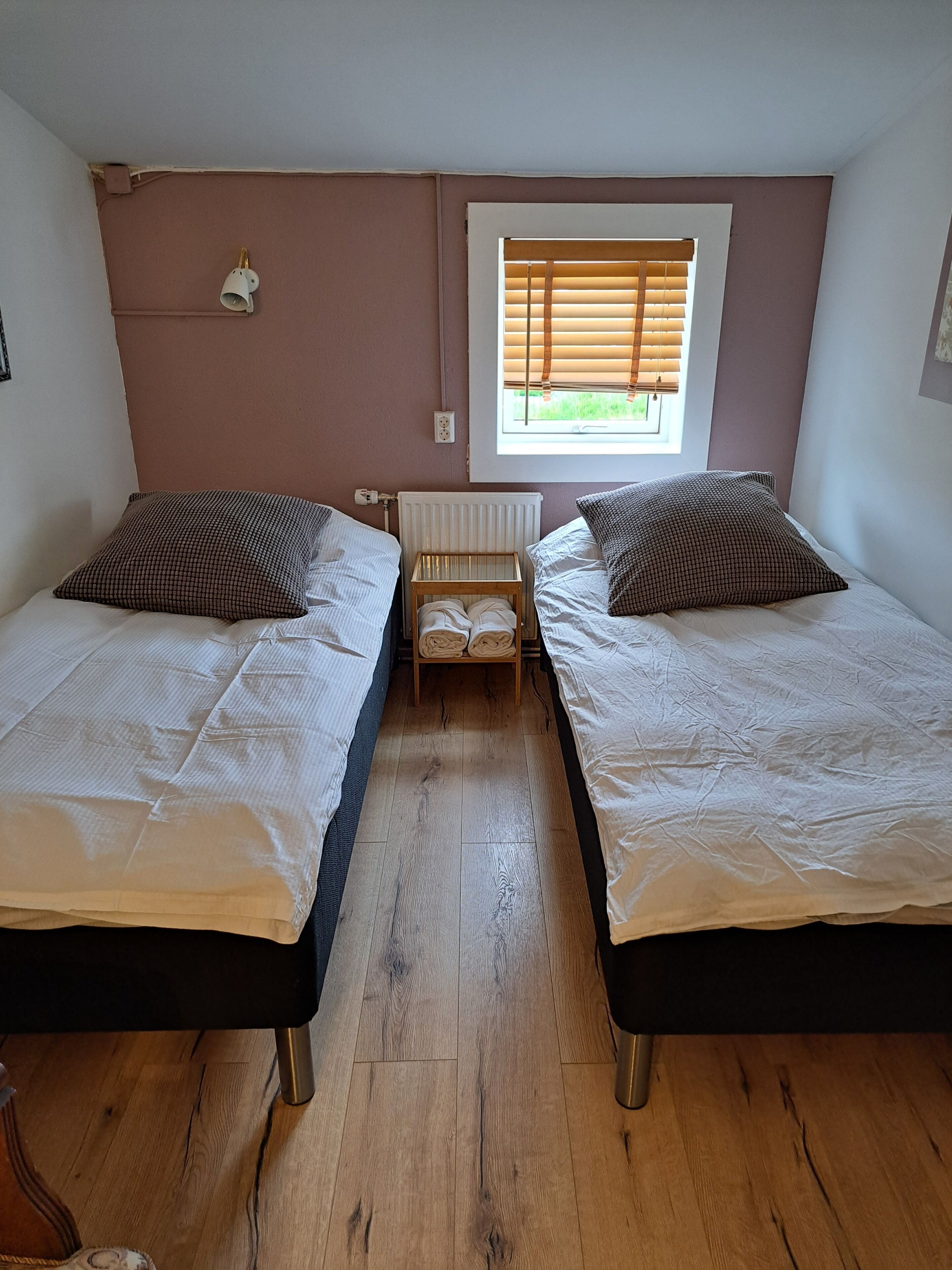 Une chambre avec deux lits, un radiateur, une fenêtre avec des stores et une petite table.