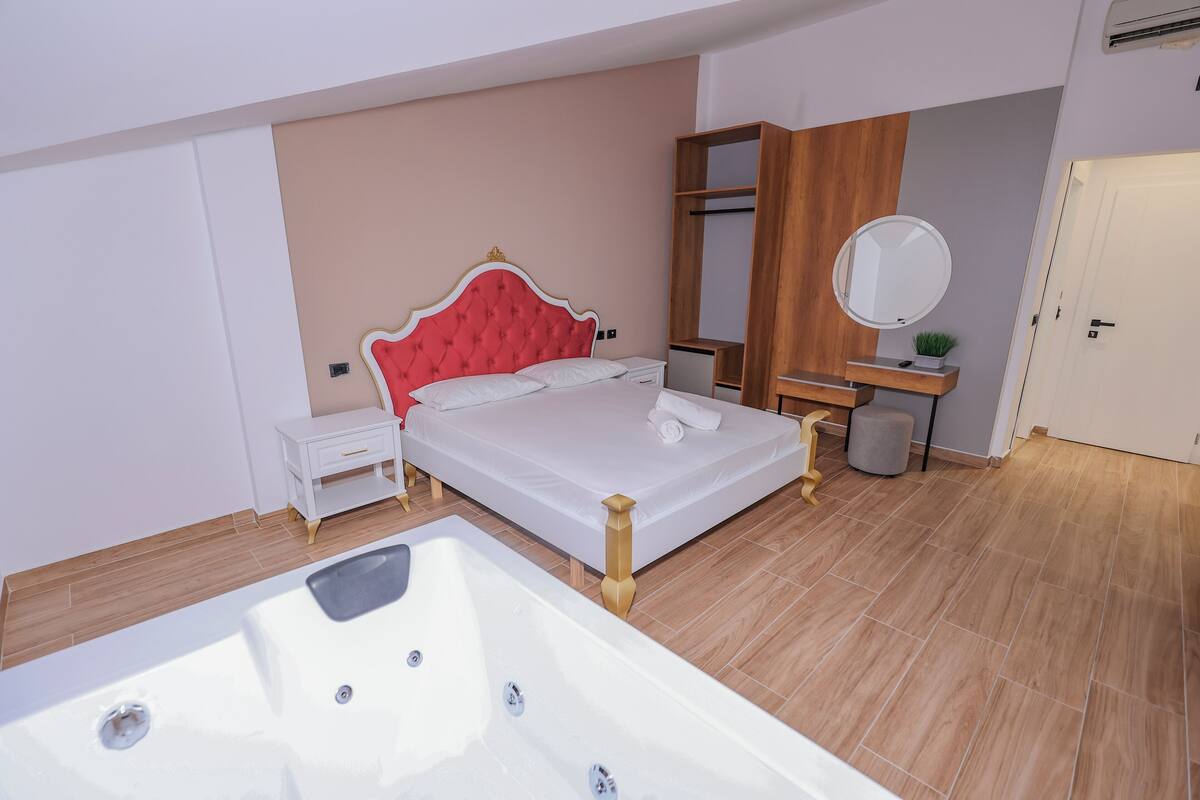 A modern bedroom with a large bed, a wooden wardrobe, a bedside table, a round mirror, and a bathtub.