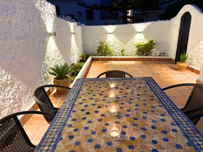 Une terrasse avec une table carrelée, des chaises, des plantes en pot et des lampes fixées au mur.