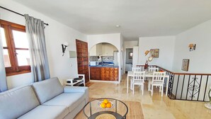 A living room with a sofa, a dining area with a table and chairs, a kitchen in the background, and a staircase leading to another level.