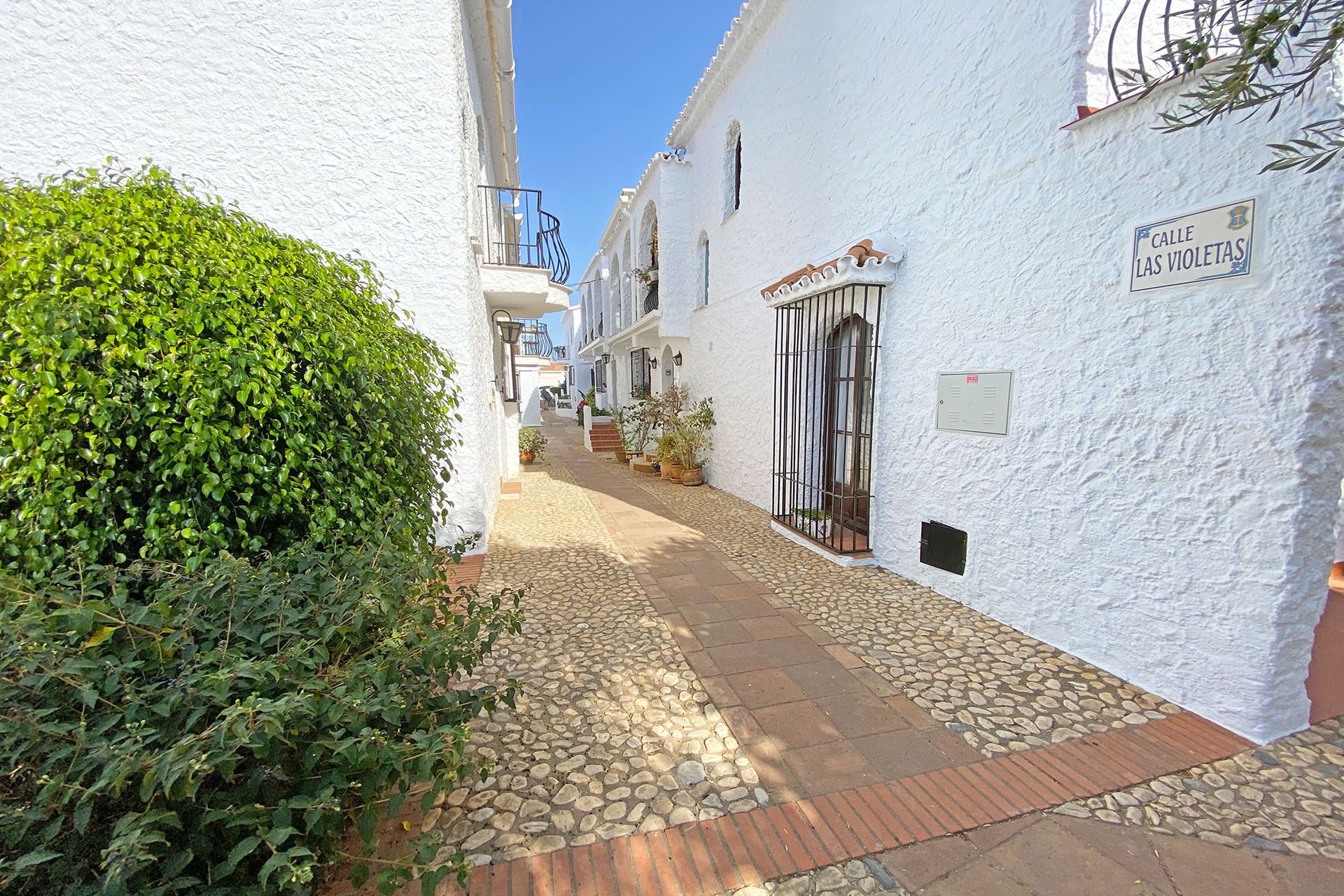 Une étroite ruelle pavée, bordée de bâtiments blancs, sur laquelle on peut lire une enseigne indiquant : « Calle de las Violetas ».