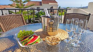 Taula amb un cubet metàl·lic, un bol de fruits secs, un plat de síndria i raïm, i diversos vasos a un balcó amb vista a la costa.