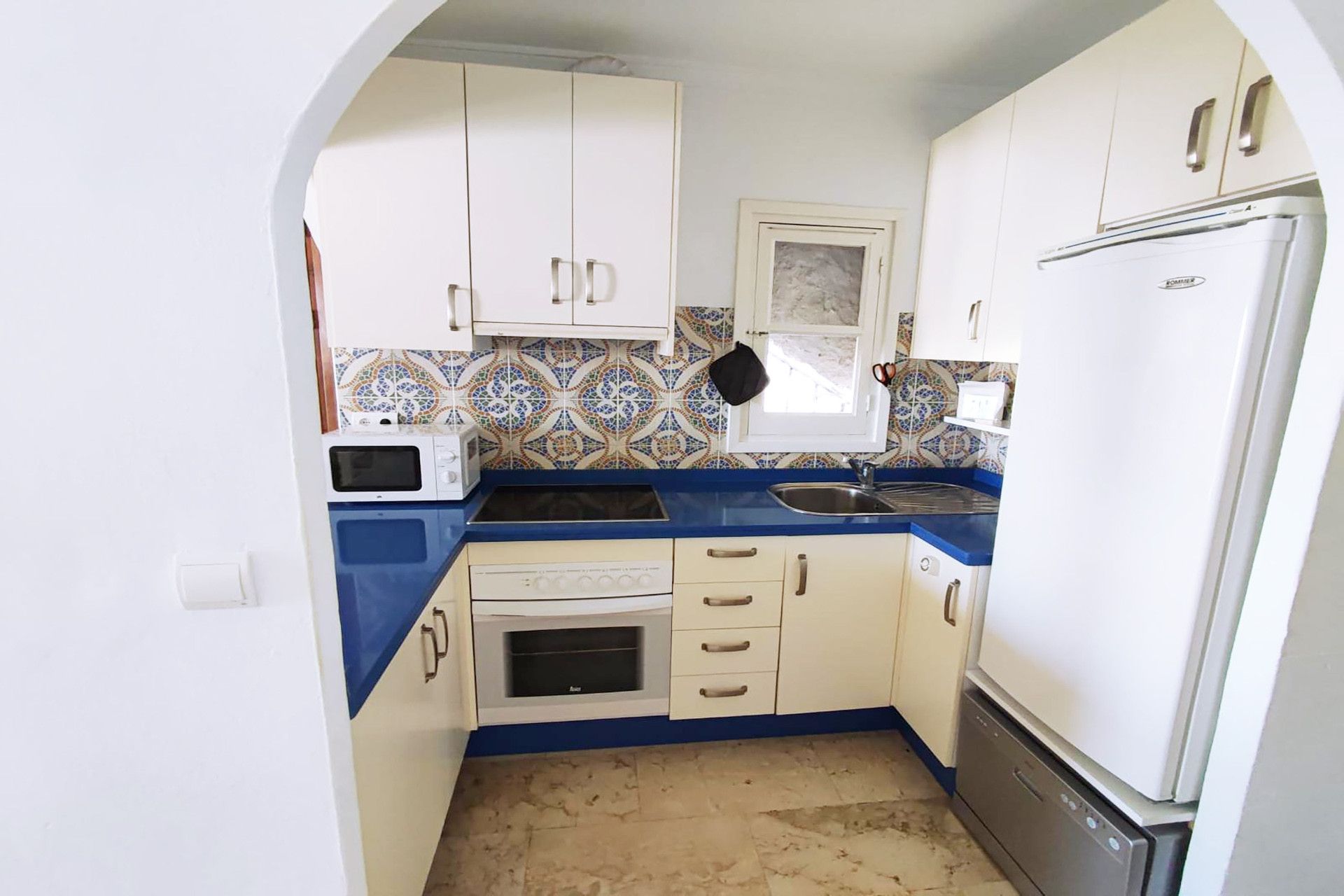 A compact kitchen with white cabinets, a blue countertop, a microwave, an oven, a sink, and a refrigerator.