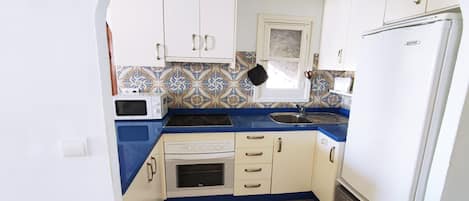 A compact kitchen with white cabinets, a blue countertop, a microwave, an oven, a sink, and a refrigerator.
