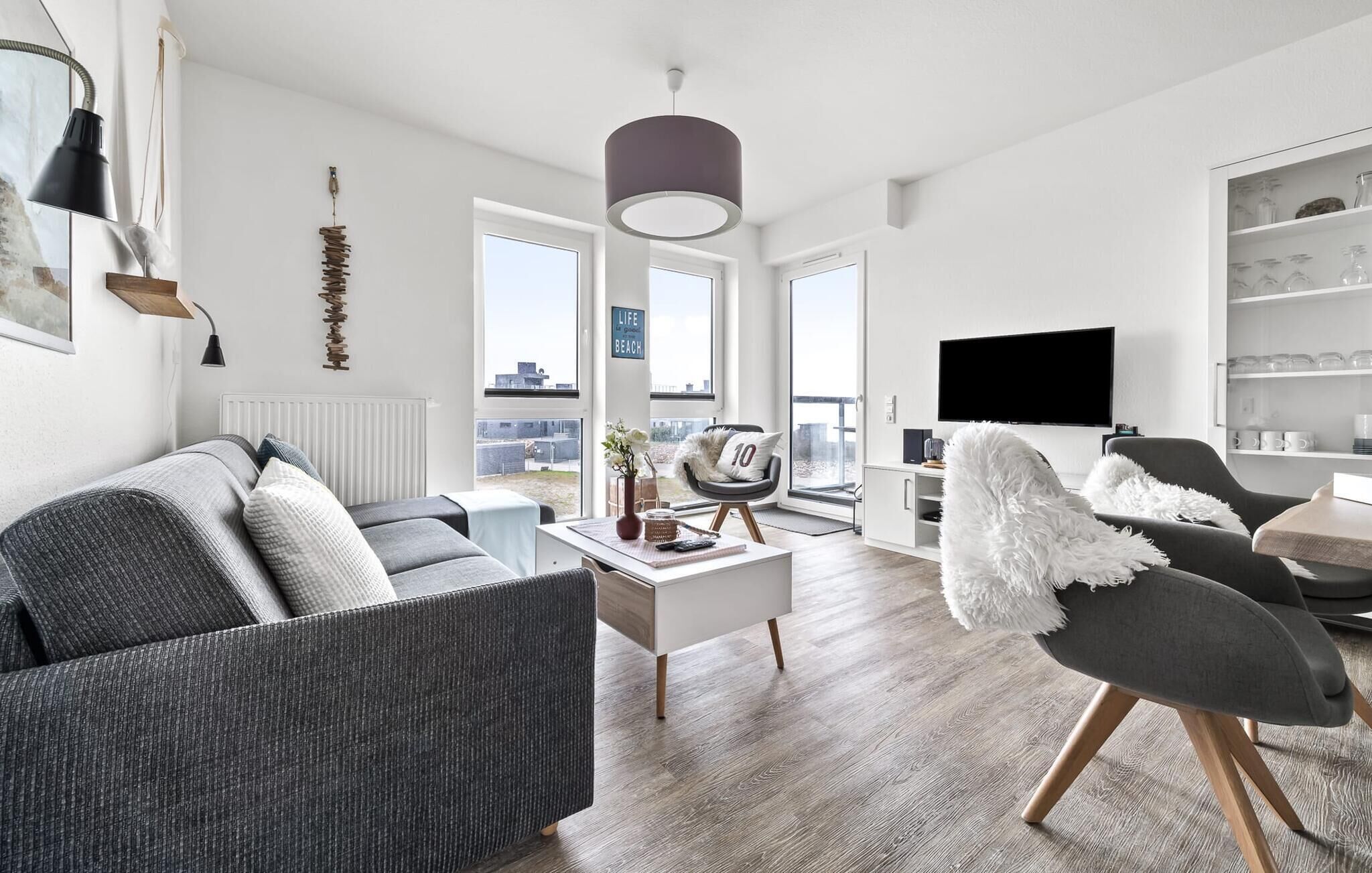 Un salon moderne avec un canapé gris, une table basse blanche et un fauteuil gris. Une grande fenêtre offre une vue sur la ville, et on y trouve également un téléviseur à écran plat et une bibliothèque où sont exposés des objets en verre.