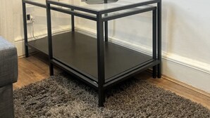 Ett bord med en glasskiva och ett svart metallstativ, placerat på en mörk matta.